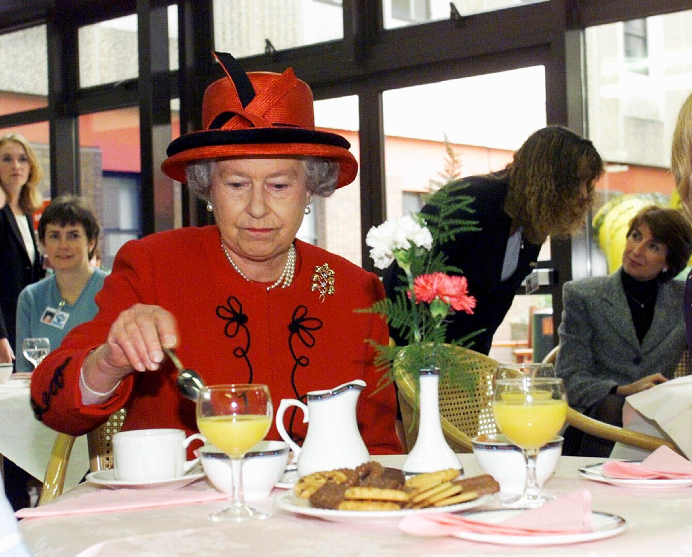 Queen Elizabeth II wears a red dress and holds a spoon inside the food house