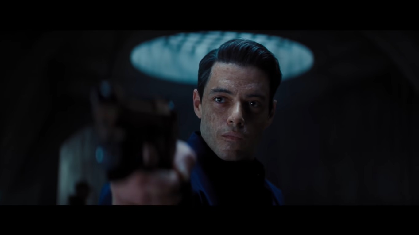 Rami Malek as Safin in ‘No Time to Die,’ with a brush-up hair and scarred face and pointing a gun