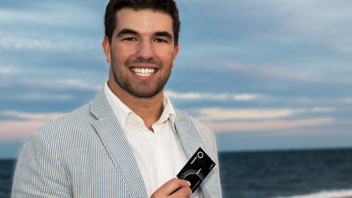 Billy McFarland in a blue striped blazer, white polo undershirt, holding a black card while smiling