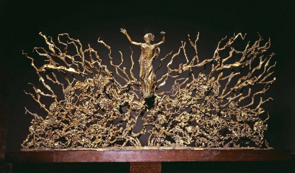 A mini golden colored metal statue of Creepy Giant Sculpture The Resurrection on a wooden shelf
