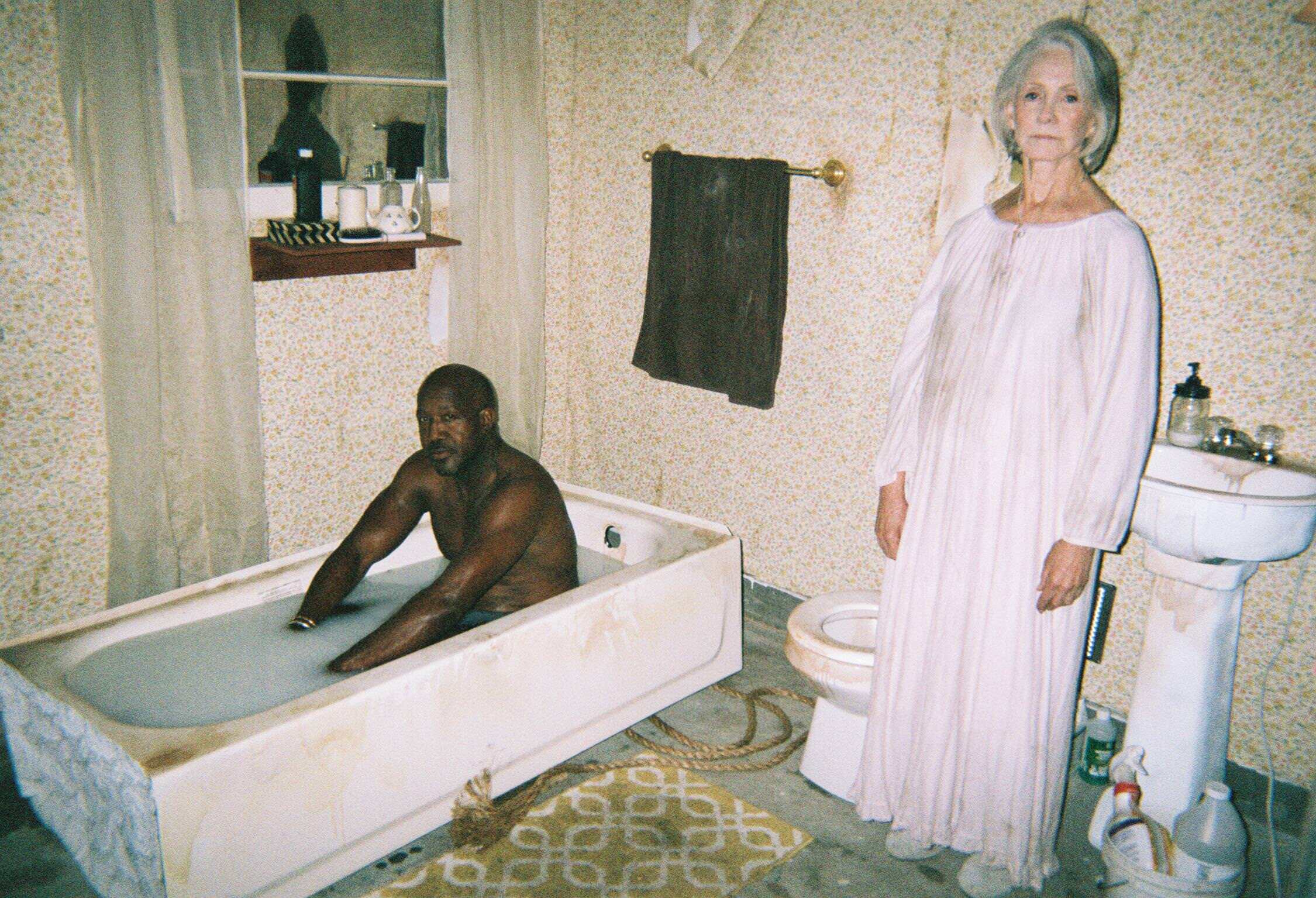 A black man in a bath tub and an old woman with stained white outfit standing beside him