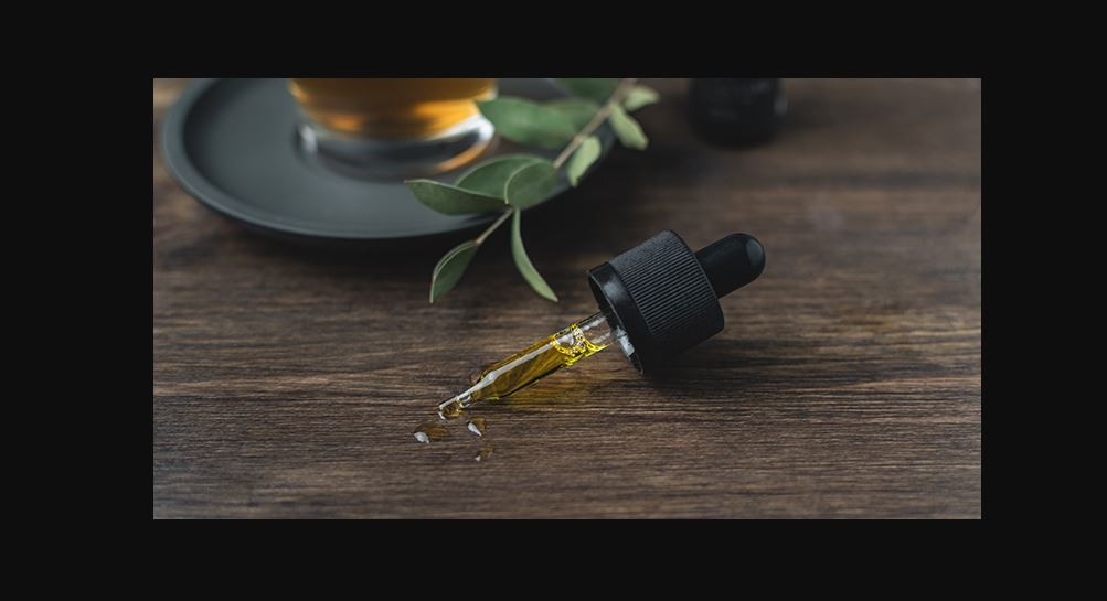 A dropper with CBD oil in it and some drops on a wooden platform it is laying