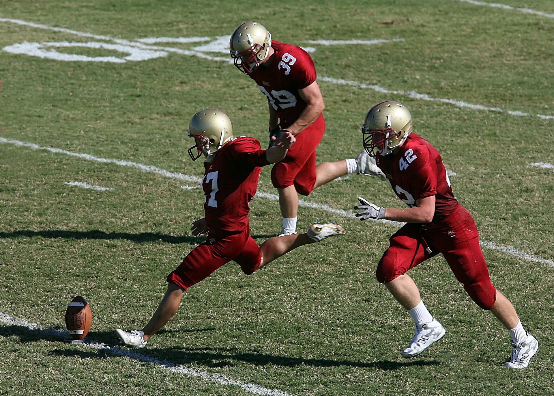 Three red uniform American football players are playing in field.