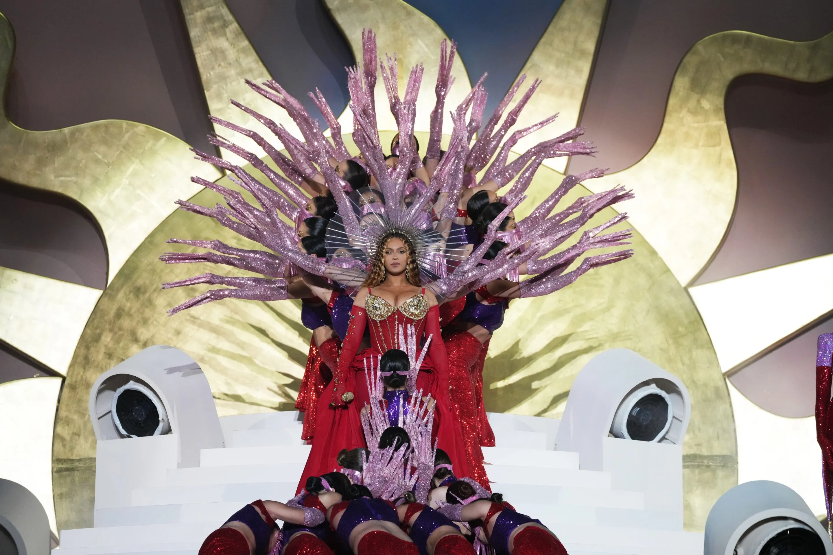 Beyoncé in an elaborate costume with a large, radiant headpiece, performing on a stage with an ornate design, evoking a regal and theatrical presence.