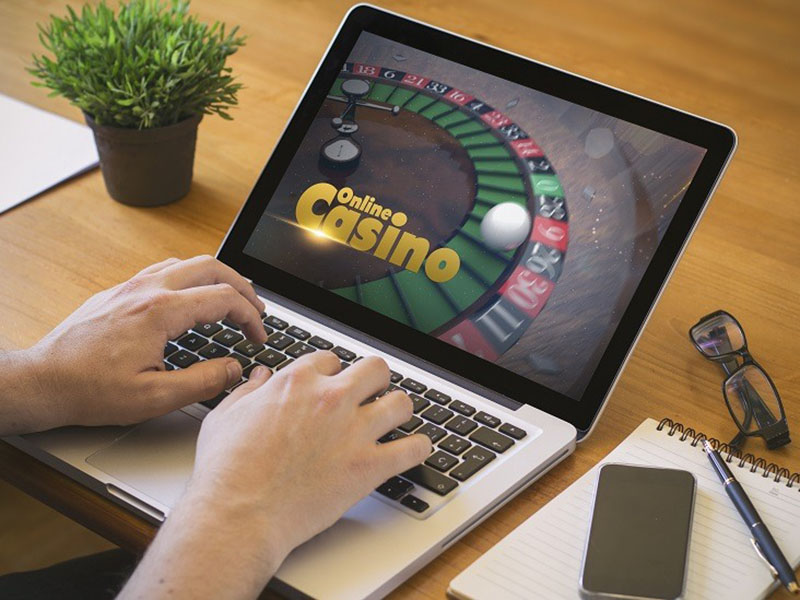 A person is using online Casino on laptop with glasses, pen, mobile phone and writing pad on the side